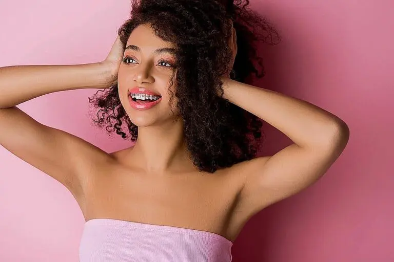 Young woman with braces at Gramercy Orthodontics in New York, NY, smiles against a pink background, hands on her head.