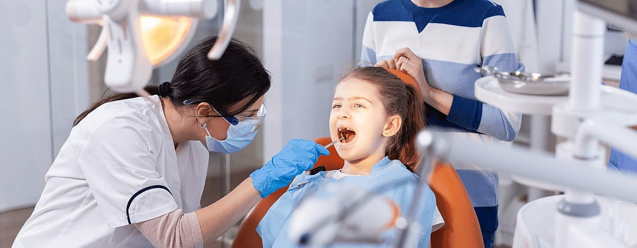 An Orthodontic Experts dentist examines a young girl in a dental chair, with her parent standing nearby. The dentist, wearing a mask and gloves, is surrounded by an array of dental equipment in NY