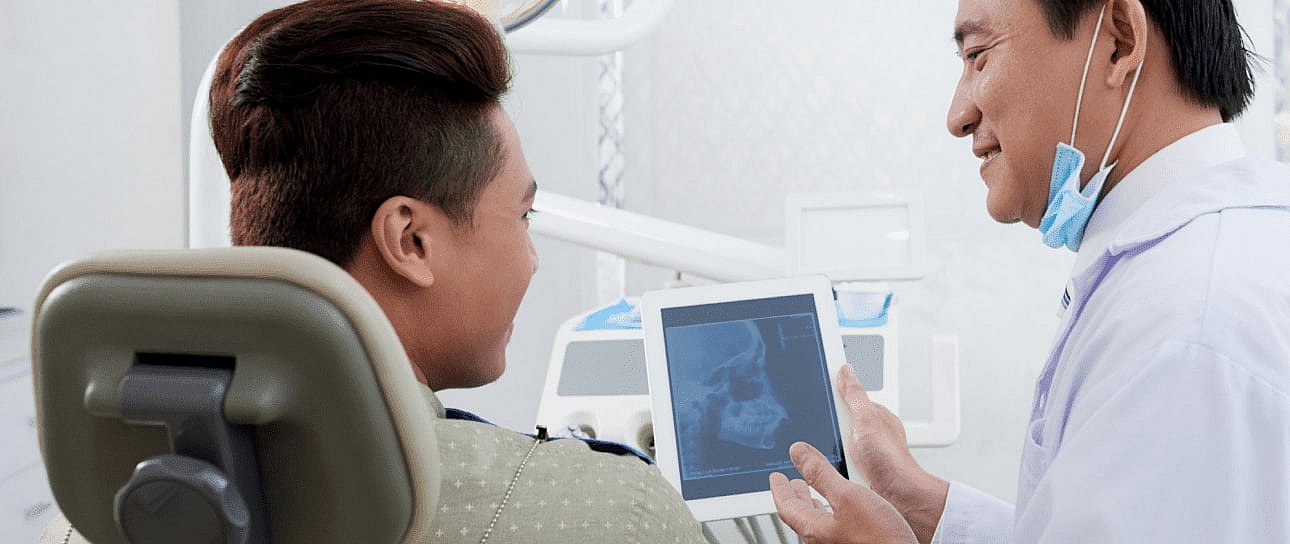 In the clinic, a dentist uses a tablet to explain a dental x-ray to a patient, highlighting areas that might require jaw surgery in NYC