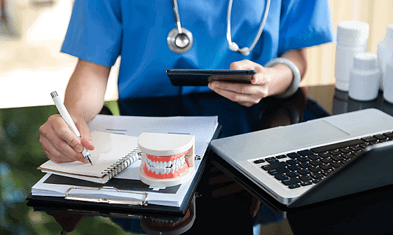 A person in scrubs writes notes beside a dental model, a laptop, and some bottles, while holding a smartphone. They glance at the screen for updates on braces insurance policies in NYC