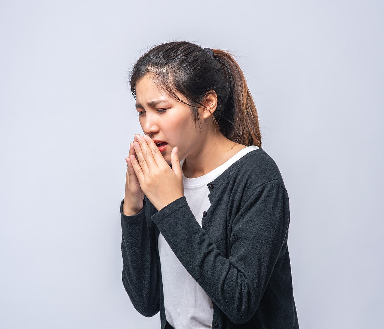 Against a light gray background, a woman in a black cardigan and white shirt sneezes into her hands, looking like she’s desperately trying not to be mistaken for a mouth breather in NYC