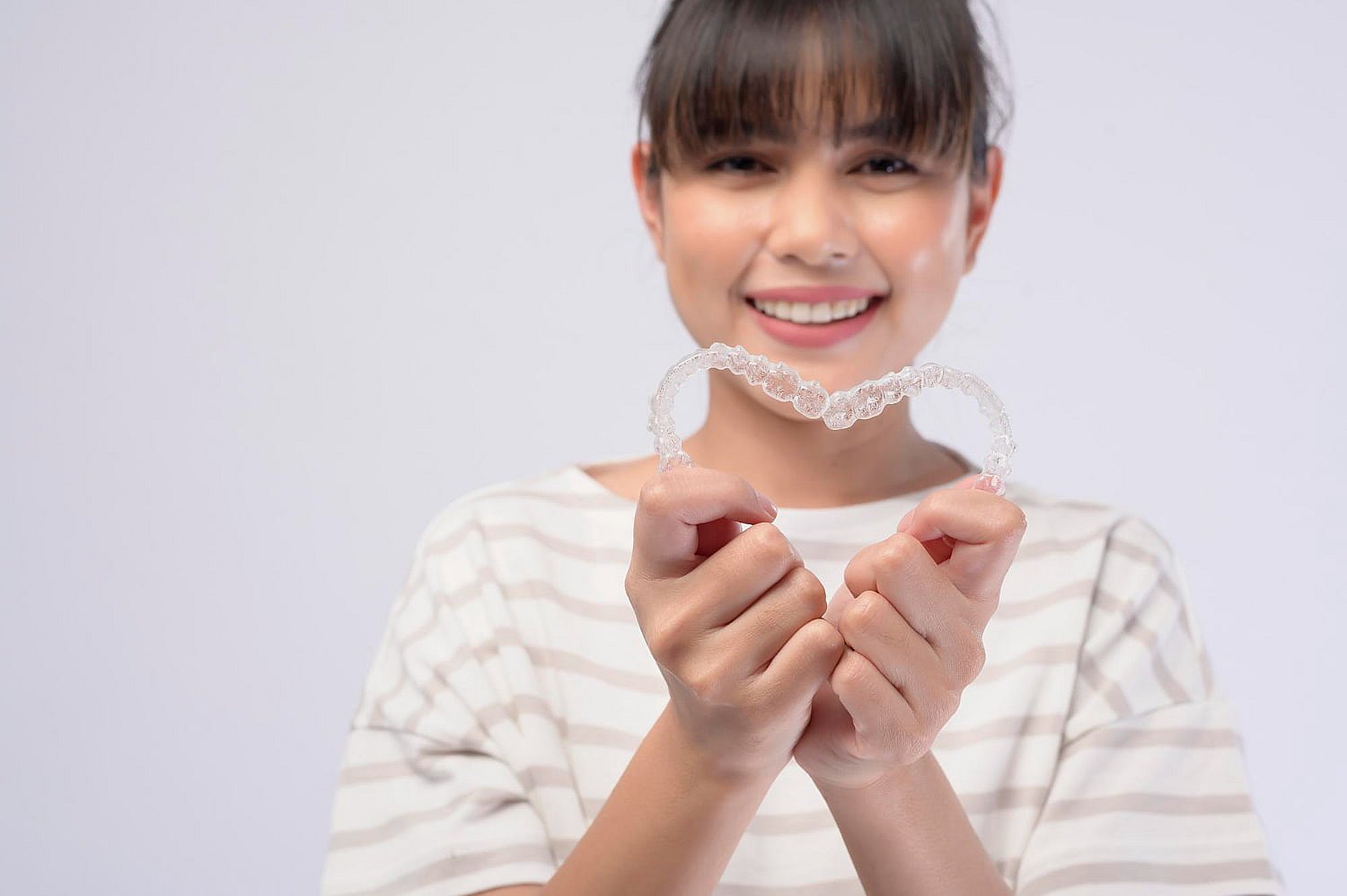 Person smiling and holding an Invisalign clear dental aligner with both hands, wearing a striped shirt against a plain background, showcasing the elegance of adult orthodontics in NYC