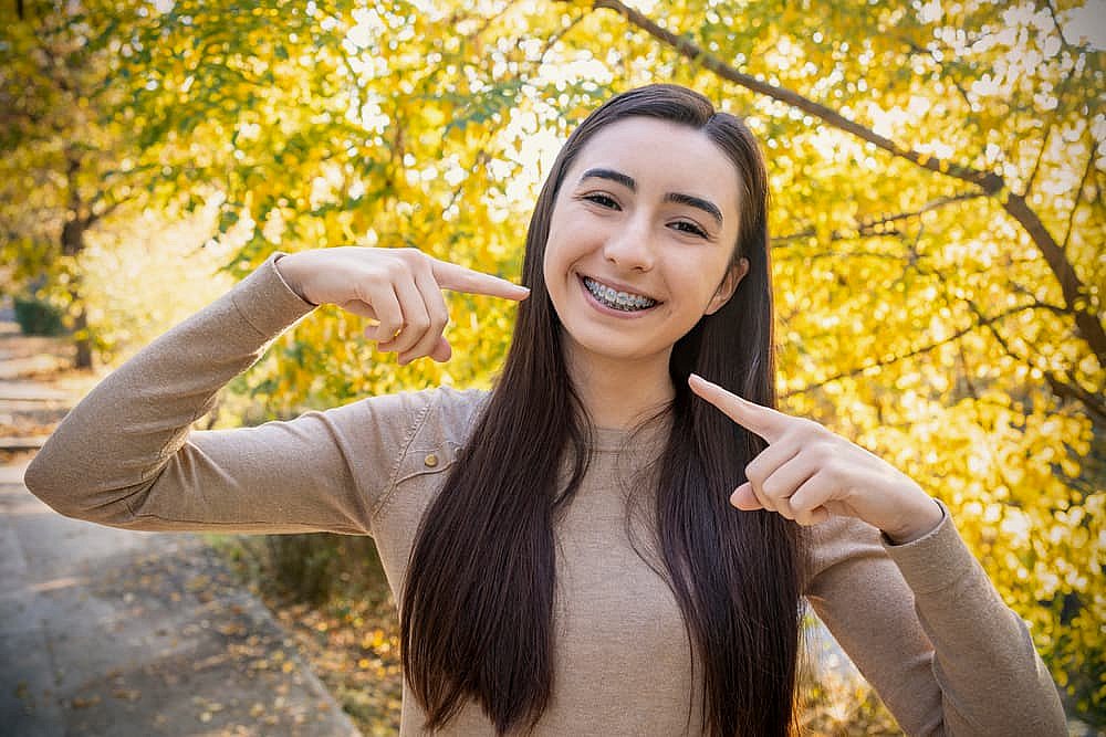 A young woman with long dark hair flashes a radiant smile, proudly pointing at her metal braces while standing in a park adorned with autumn trees in NYC