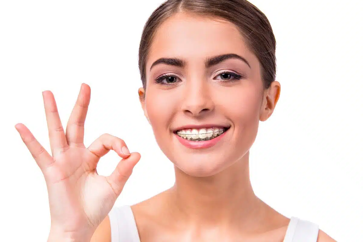 A person with ceramic braces beams a confident smile, making an "OK" hand gesture against a white background in NYC