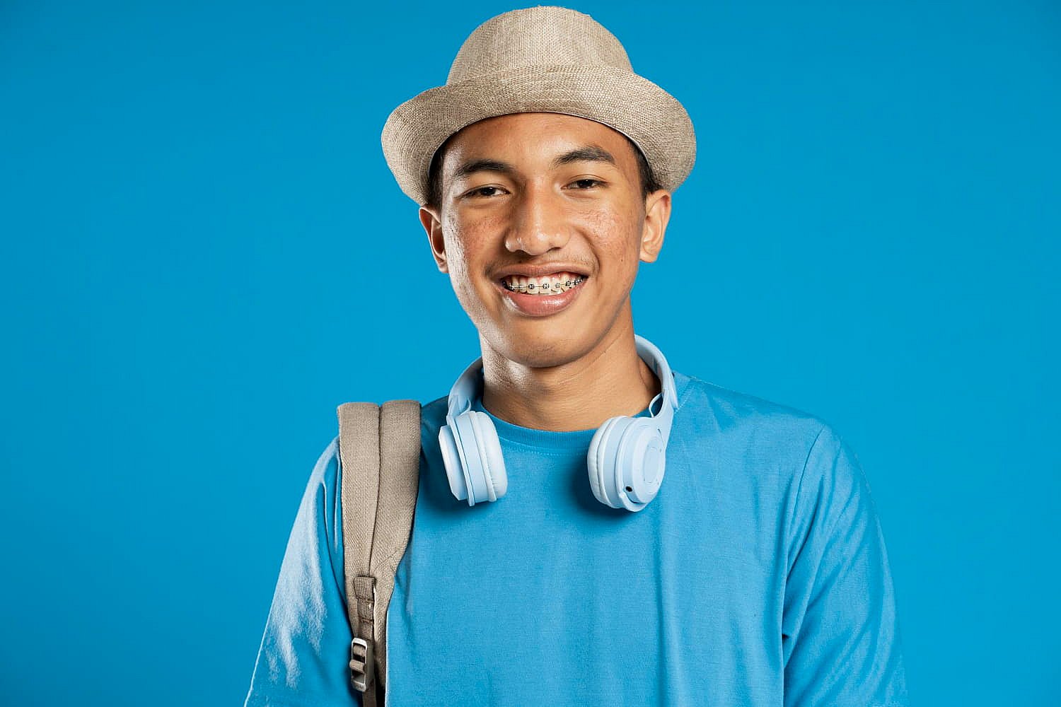 A young person in a straw hat and blue shirt beams with confidence, their metal braces glinting under the light. White headphones rest around their neck, and a backpack is slung over one shoulder, all set against a serene blue background in NYC