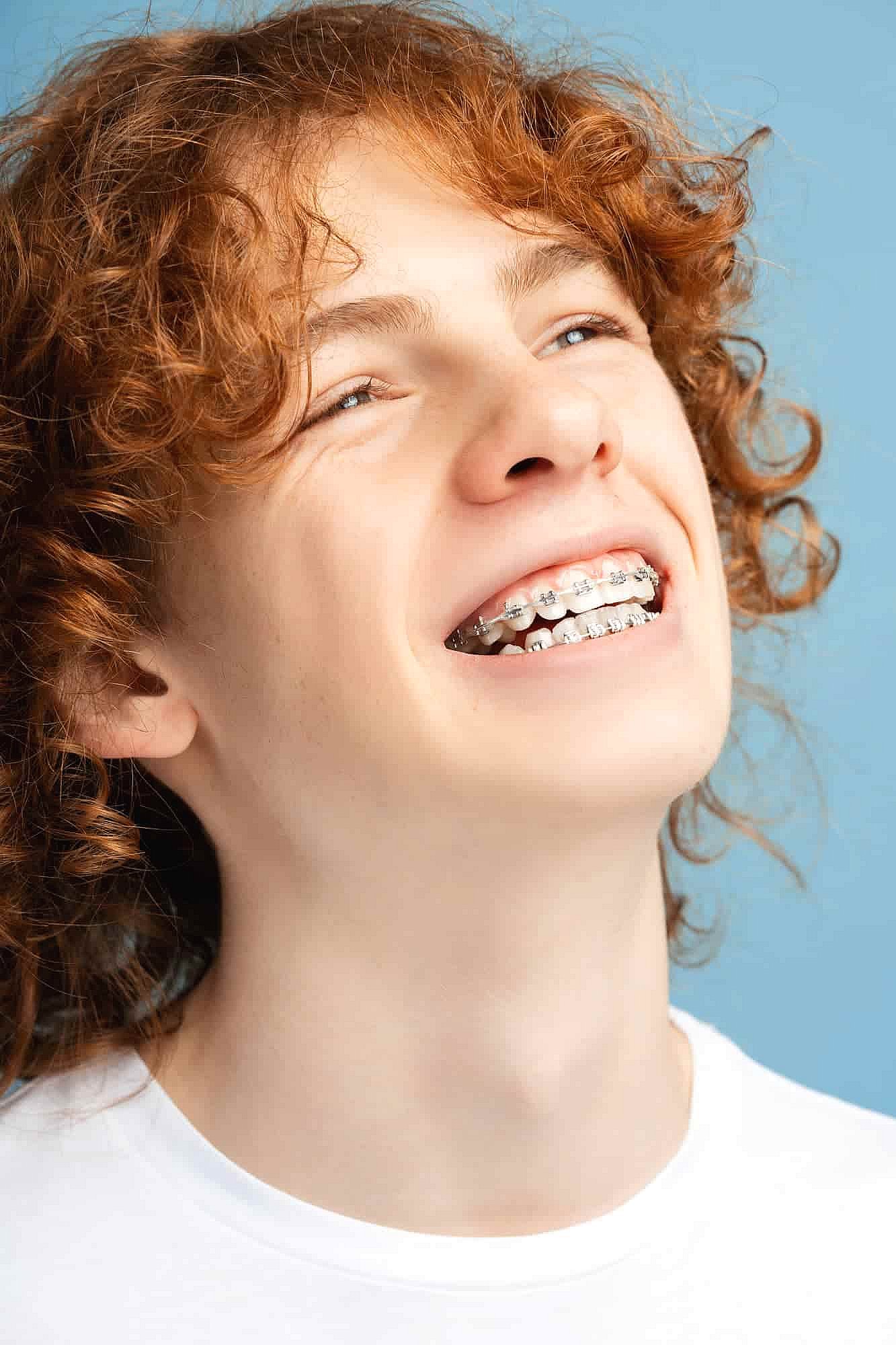 A smiling person with curly red hair and braces, showcasing the wonders of adult orthodontics, wears a white shirt against a serene blue background in NYC