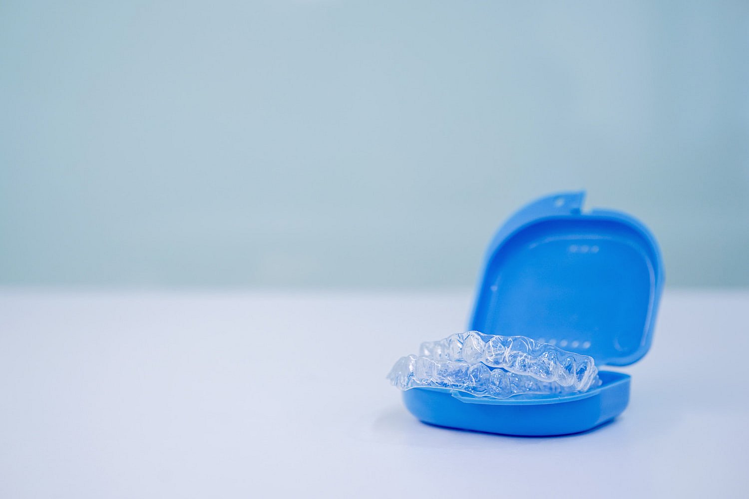 Clear dental aligners rest in an open blue case on a pristine white surface, set against a serene light blue background. It's essential to research Invisalign costs as you consider this discreet solution for a confident smile in NYC
