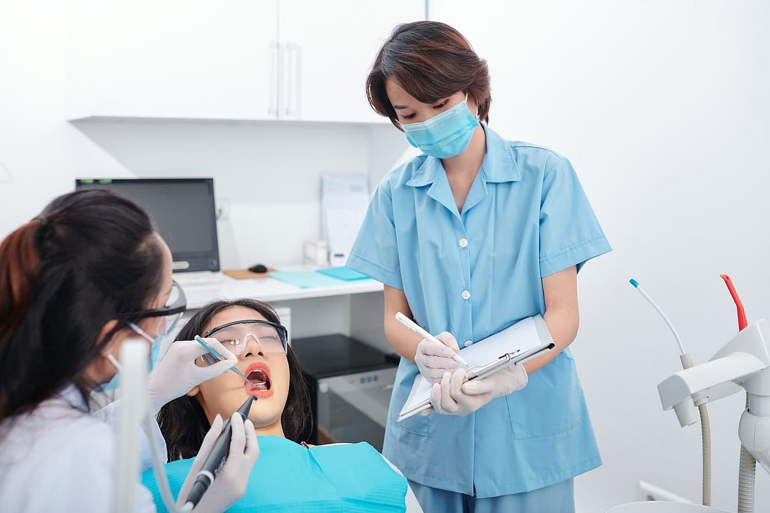 A dentist examines a patient's teeth, discussing adult orthodontics options, while an assistant takes notes in the dental office in NYC
