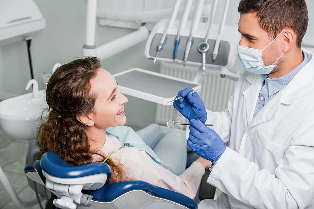 A dentist wearing gloves and a mask examines a smiling patient with metal braces in a dental chair, holding dental tools in NYC