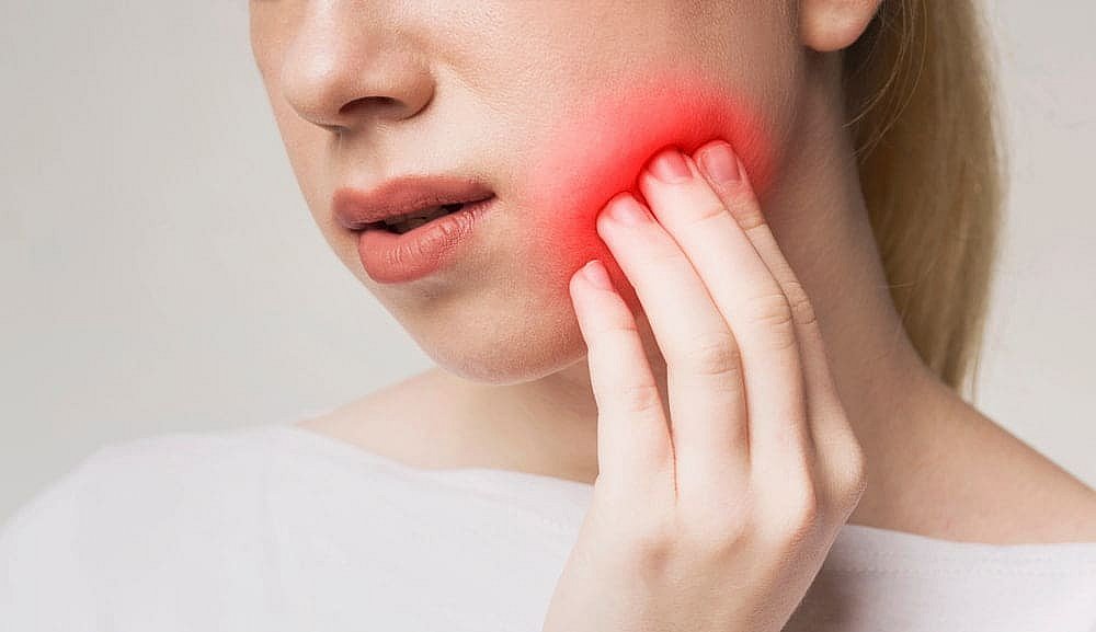 A person touches their cheek, revealing a red glow that hints at pain or discomfort—perhaps pondering the decision of Invisalign vs Braces and its potential impact in NYC
