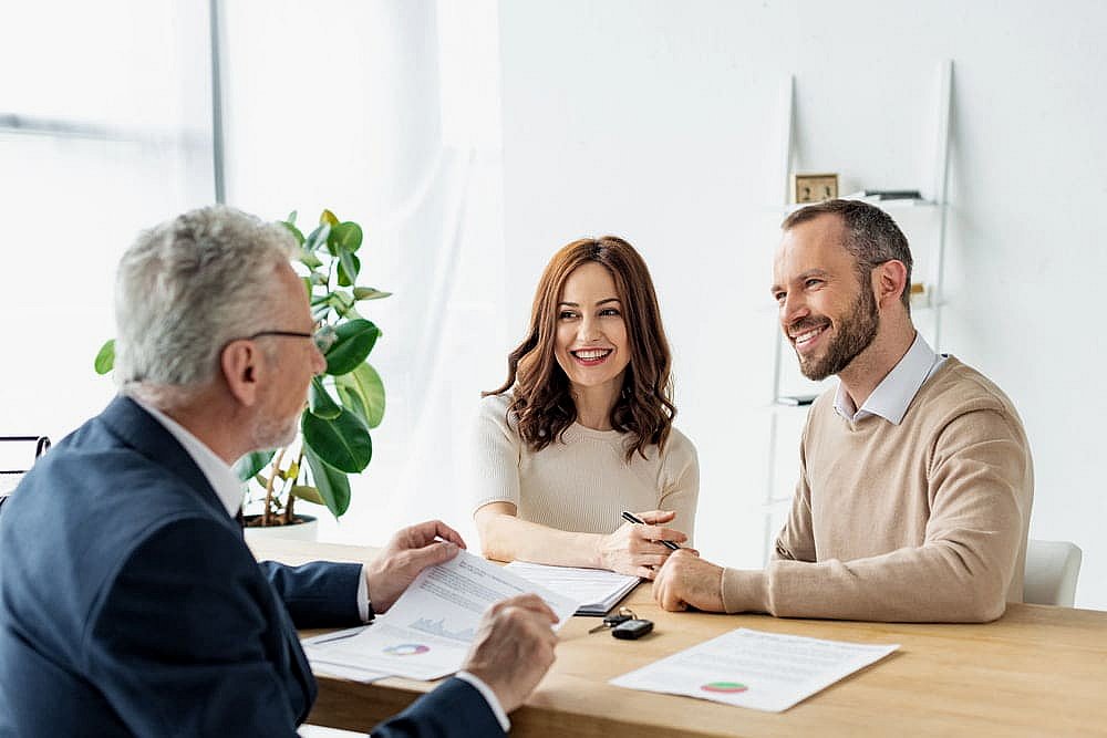 In an office setting, a couple discusses an Invisalign payment plan with an older man at the table. Surrounding them are lush green plants and a pristine white bookshelf, creating a serene backdrop for their conversation about future smiles in NYC