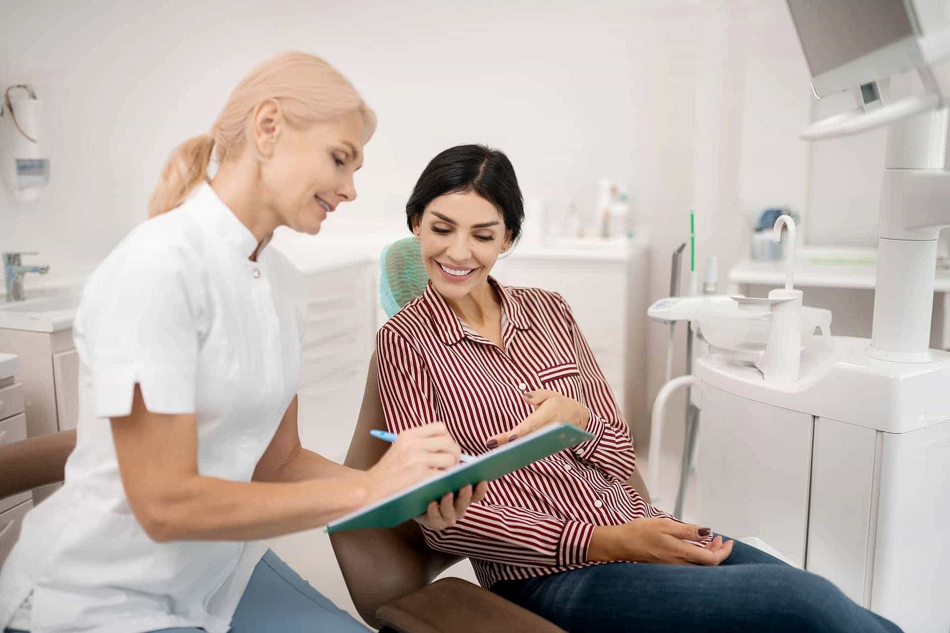 A dental professional discusses a document with a patient sitting in a dental chair, both smiling about the affordable Invisalign options available to enhance their smile in NYC