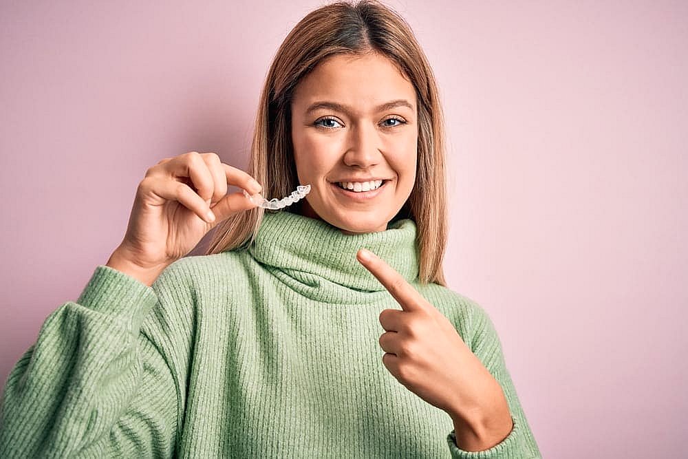 Person in a green sweater smiles while holding and pointing to a clear dental aligner against a pink background, showcasing the benefits of an Invisalign payment plan in NYC
