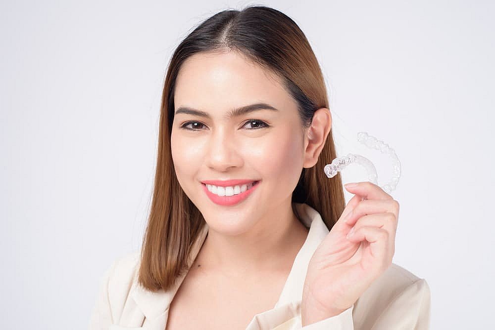 A person smiling while holding a clear Invisalign aligner in their hand, wearing a light-colored jacket against a white background, possibly wondering if this investment in their smile will be covered by insurance in NYC