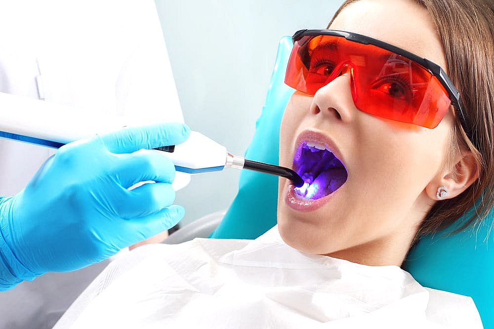 A patient wearing protective eyewear receives dental treatment with a UV light device while lying back in the dentist's chair, addressing concerns like tongue thrust in NYC