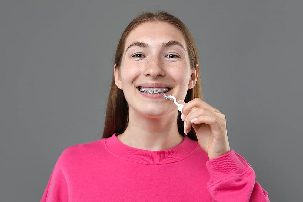 A young person with long hair, wearing a pink sweater, is smiling while using a dental flosser on their braces against a gray background—a perfect snapshot in the ongoing conversation of Invisalign vs. Braces in NYC