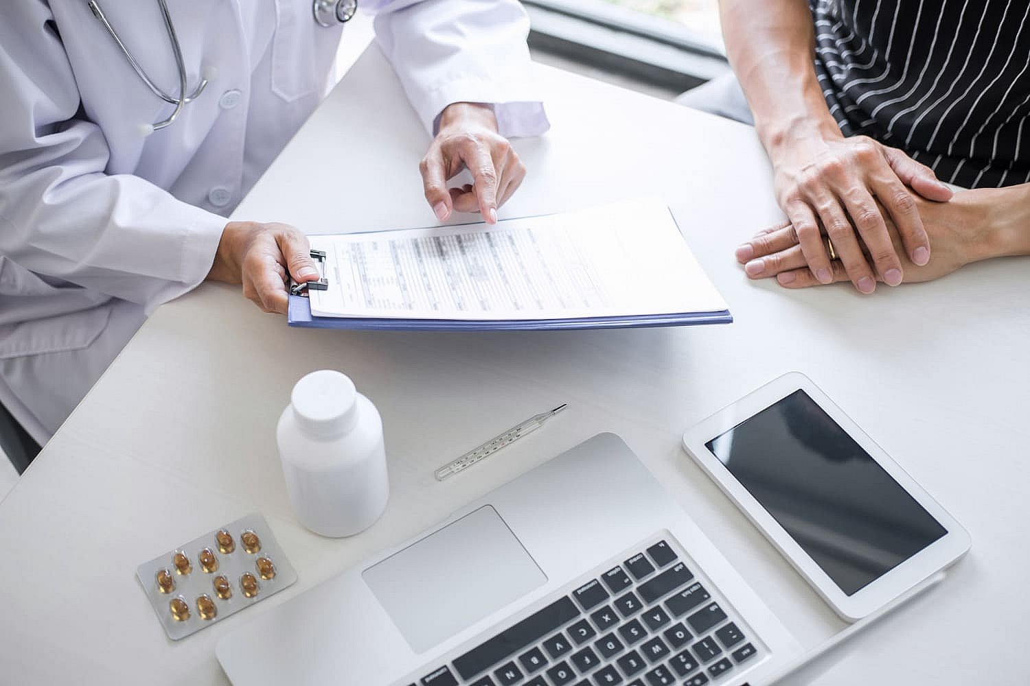 Doctor and patient reviewing a document at a desk equipped with a laptop, tablet, medicine bottle, pills, and discussing options like a braces payment plan in NYC