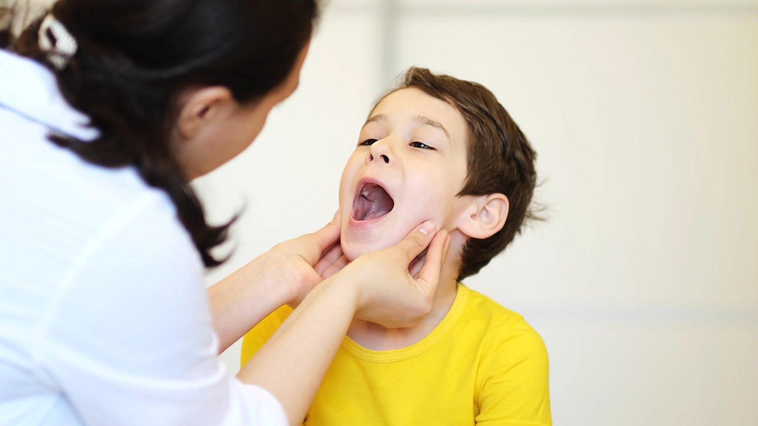 A child in a yellow shirt sits with their mouth open while an adult, perhaps pondering Invisalign vs braces for the future, examines their throat, gently holding the child's chin and jaw in NYC