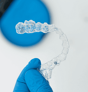 A hand in a blue glove holds a transparent dental aligner against a blurred blue and white background, highlighting the benefits of modern orthodontics with Invisalign payment plans available for accessible and flexible treatments in NYC