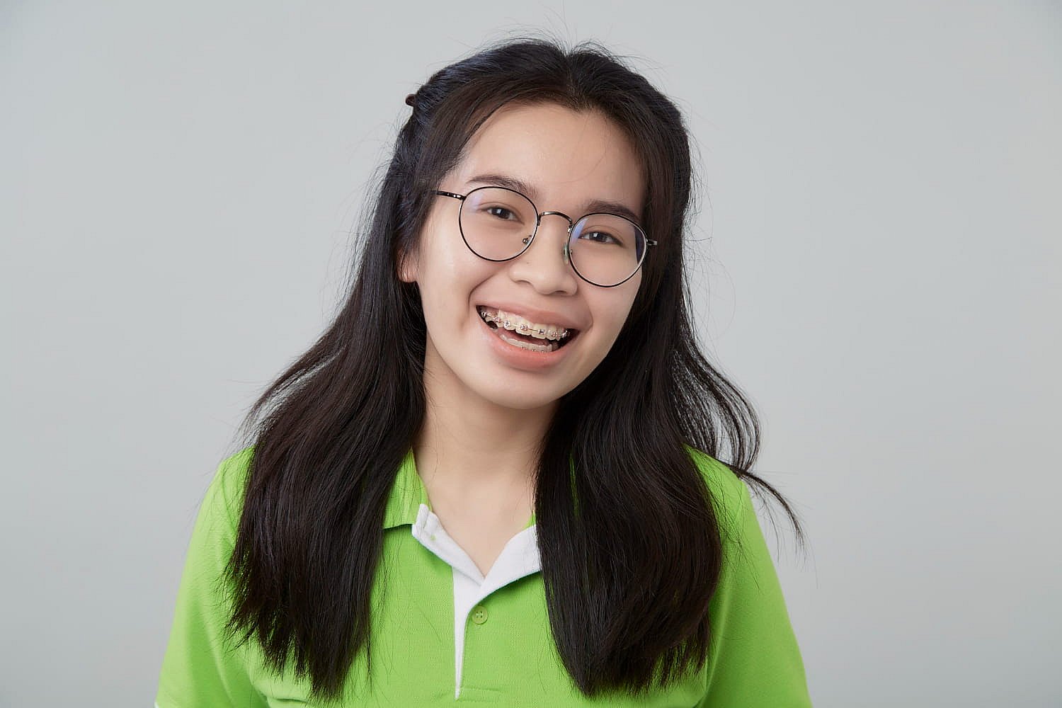 A person with long dark hair and glasses flashes a warm smile, showcasing metal braces while wearing a green polo shirt in NYC