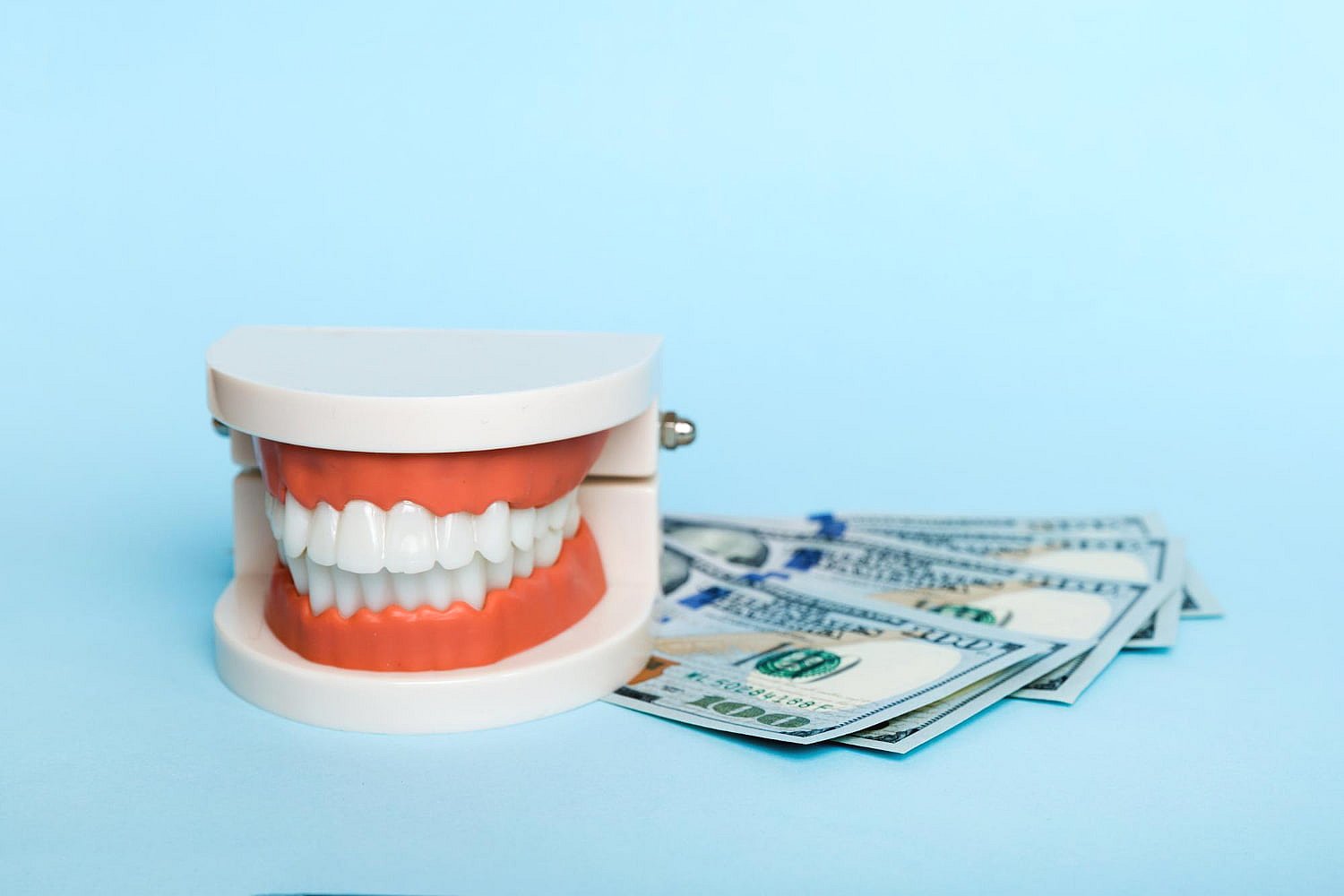 A dental model with a set of teeth, complete with braces, is placed beside several scattered U.S. hundred-dollar bills against a light blue background, subtly hinting at the cost and value of proper dental insurance in NYC