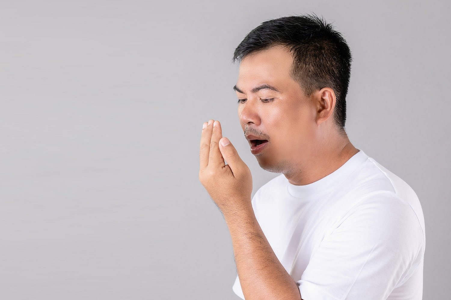 A man in a white shirt gazes at his hand while discreetly covering his mouth, perhaps checking for an underbite, set against a plain gray background in NYC