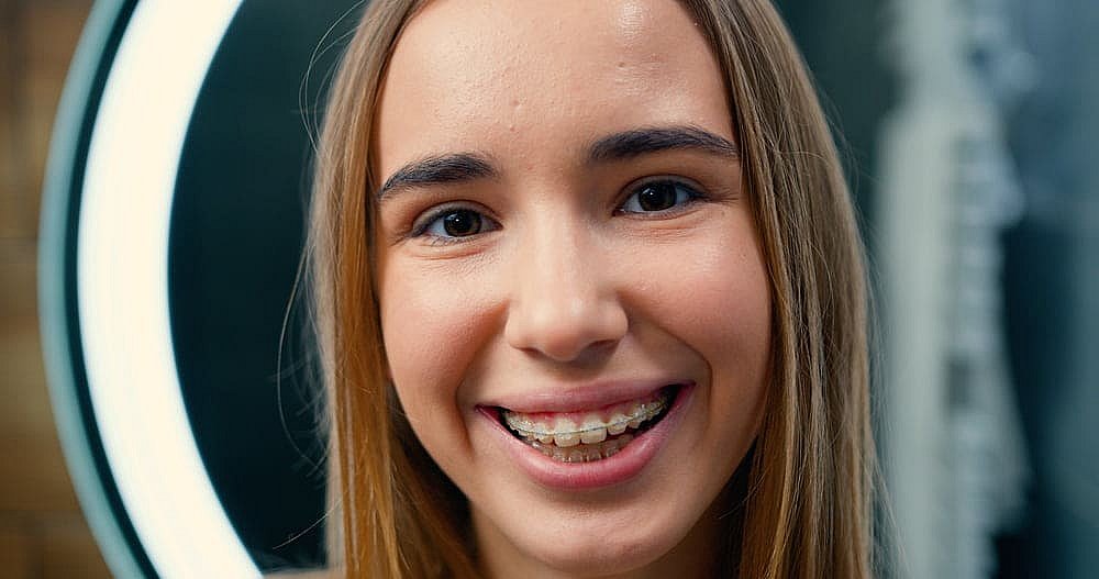 Teen girl with long hair smiles, proudly showing her clear braces. A circular light is visible in the background in NYC