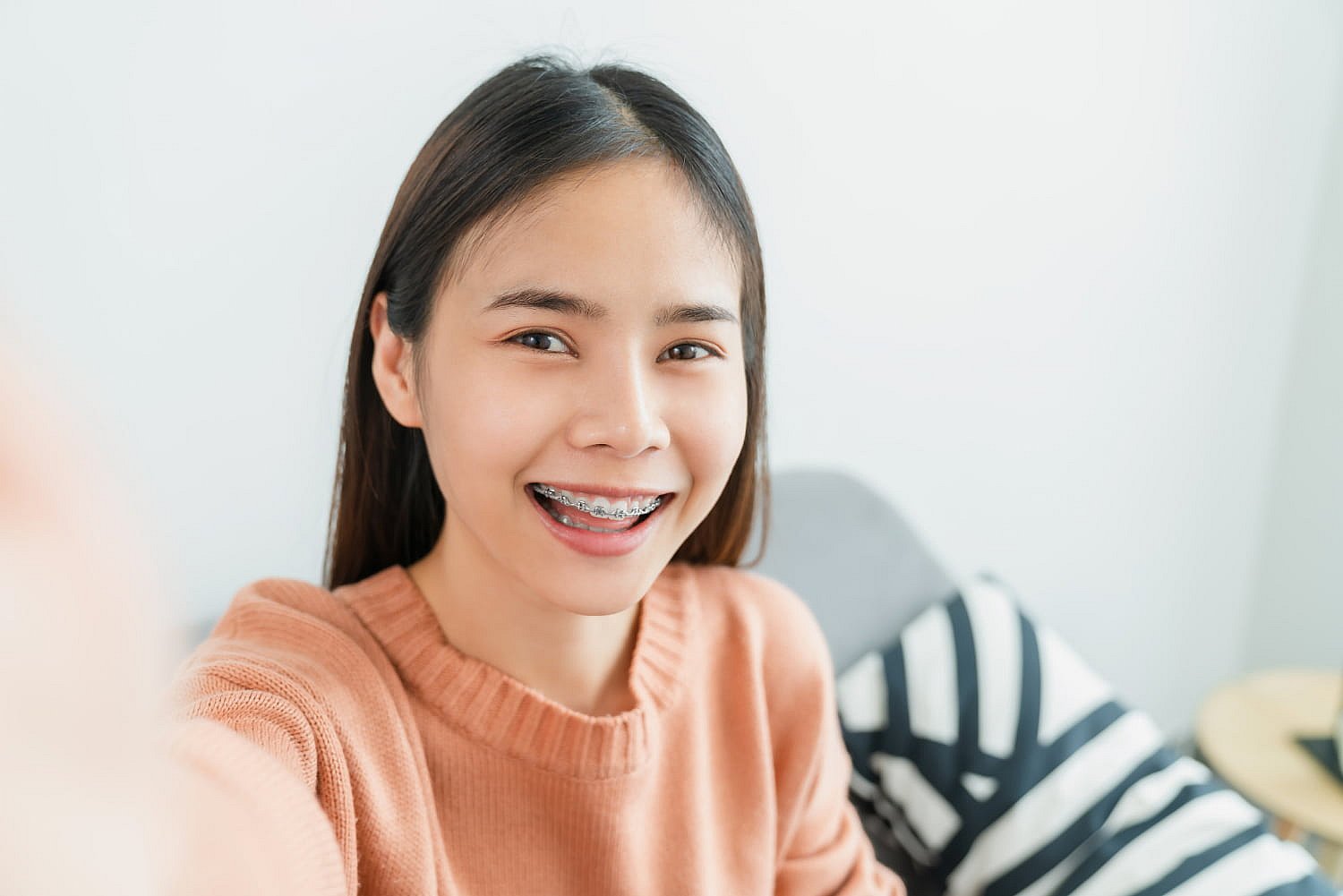 A smiling woman with long hair, showcasing her newly acquired braces, takes a selfie indoors in her peach sweater. The striped pillow adds a cozy touch to the scene, hinting at the investment behind her radiant smile and the braces cost worth every penny in NYC