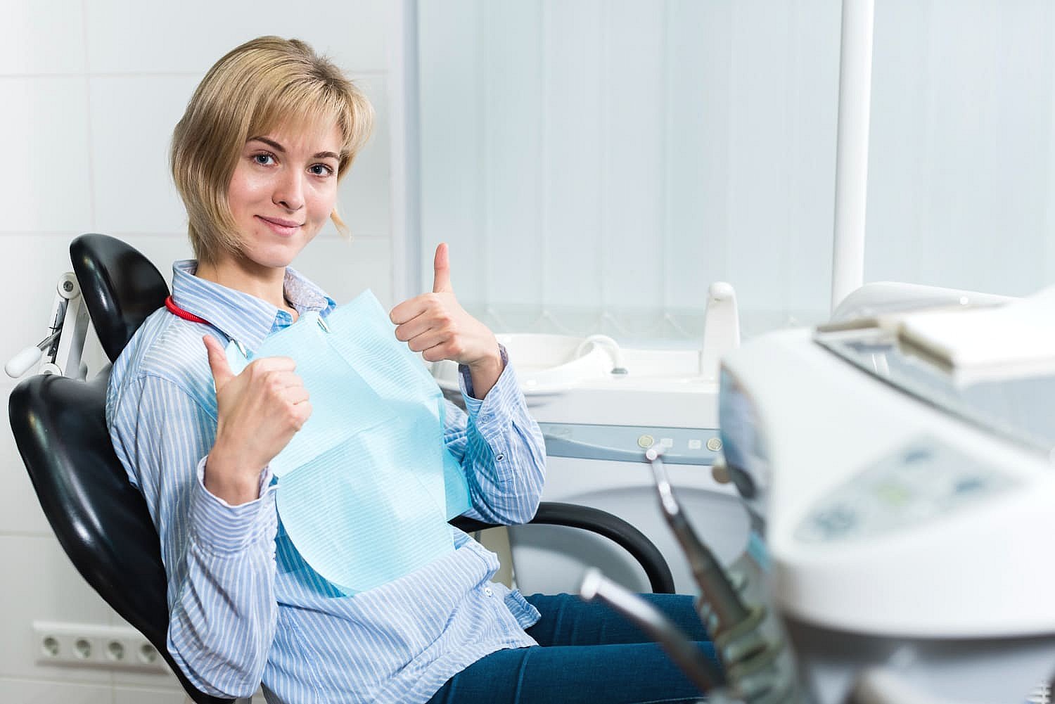 A person sits in a dental chair, wearing a blue bib and giving two thumbs up with a bright smile, confident knowing their braces are covered by insurance. Dental equipment is neatly arranged in the foreground in NYC