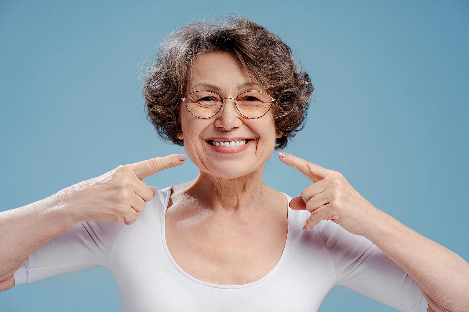 An elderly woman with glasses is smiling warmly and pointing at her teeth, subtly showcasing the confidence that comes with good braces. She's wearing a crisp white shirt against a calming blue background in NYC