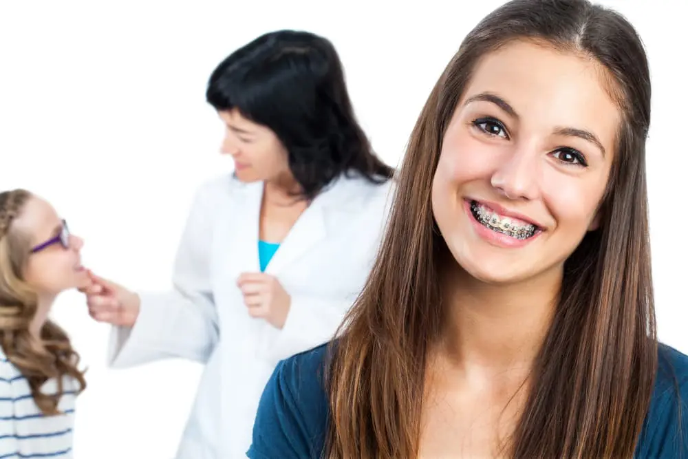 A teen with braces, contemplating the cost, smiles at the camera while a woman in a white coat checks another girl's teeth in the background in NYC