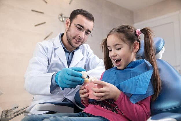 A children's orthodontist shows a young girl a dental model. She looks excited, sitting in the dental chair with her protective bib, eager to learn about orthodontic appliances in NYC