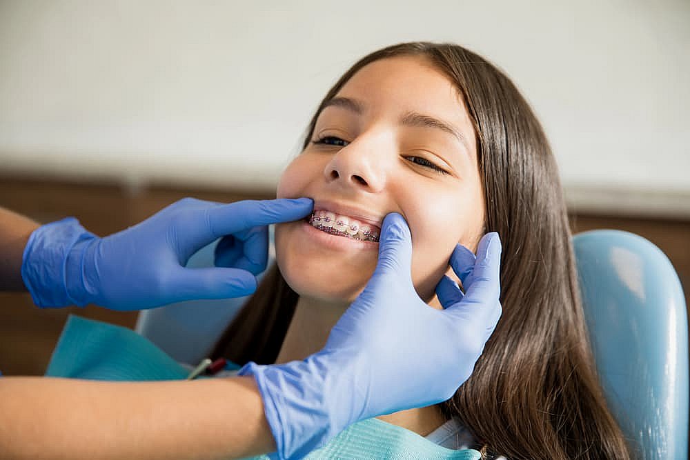 A girl with long hair and braces is having her teeth examined by a dentist wearing blue gloves, all while pondering the cost of maintaining her sparkling smile in NYC