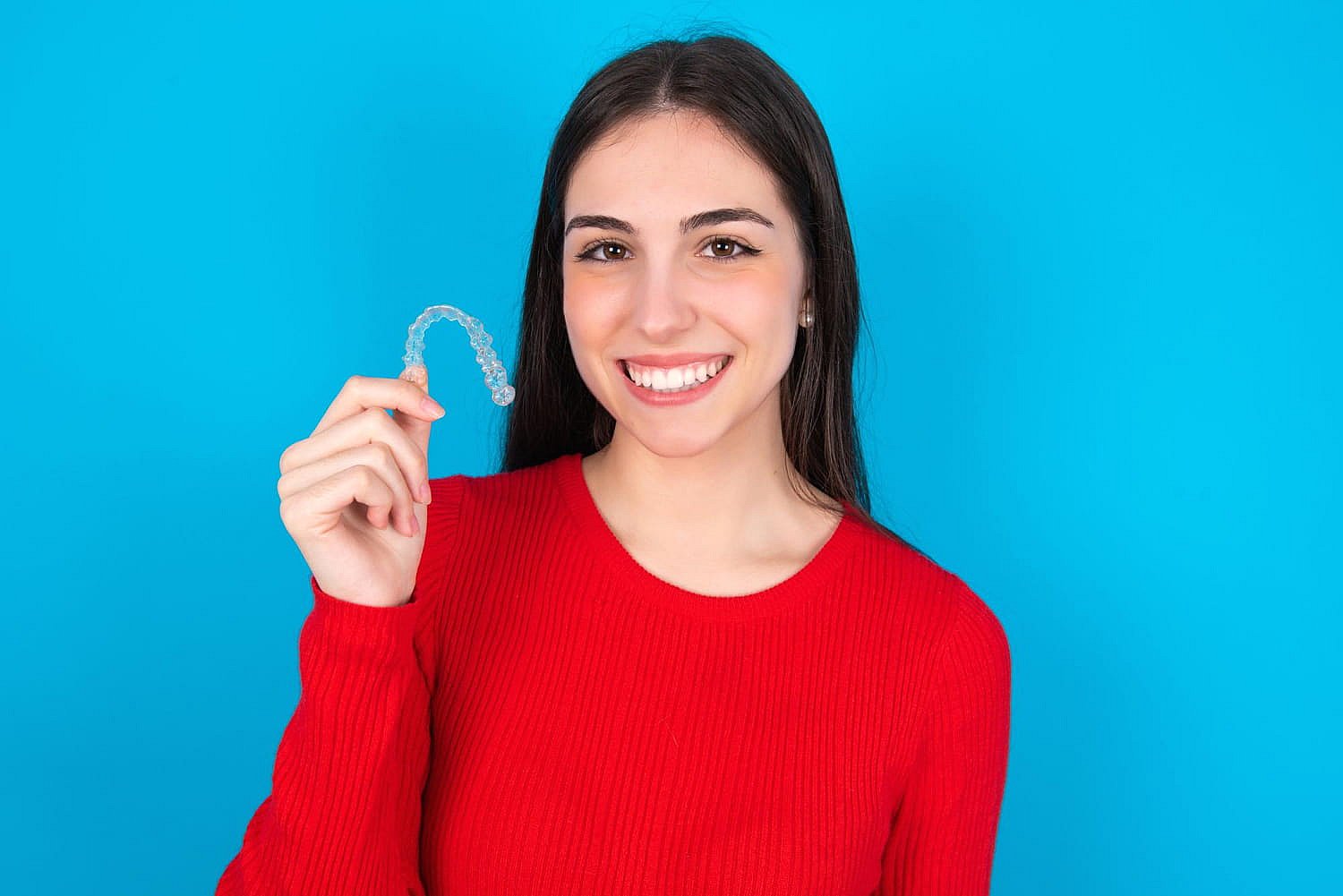 A woman in a red sweater beams as she holds a clear dental aligner against a blue background, contemplating if Invisalign might be covered by insurance in NYC