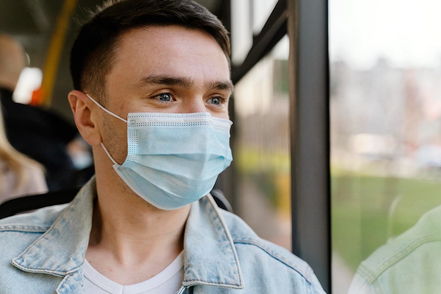 A person wearing a blue face mask and denim jacket gazes pensively out of the bus window, mindful not to be mistaken for a mouth breather in NYC