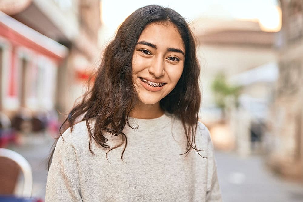 A young woman with long hair and a radiant smile showcases her metal braces, standing outdoors on a street with blurred buildings in the background in NYC