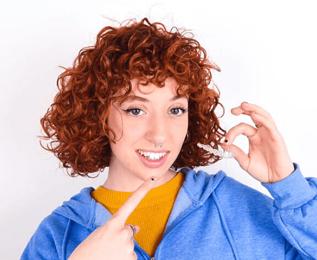 A person with curly red hair in a blue hoodie is smiling and pointing at their Invisalign dental aligner in NYC
