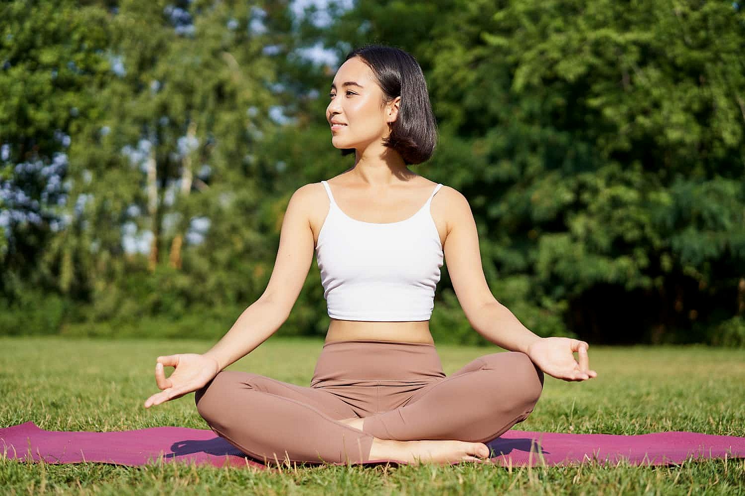 A person sits cross-legged on a yoga mat in a park, wearing a white tank top and beige pants. Amidst the tranquil trees in the background, they focus on their breathwork, ensuring they're no mere mouth breather but fully engaged in mindful respiration in NYC