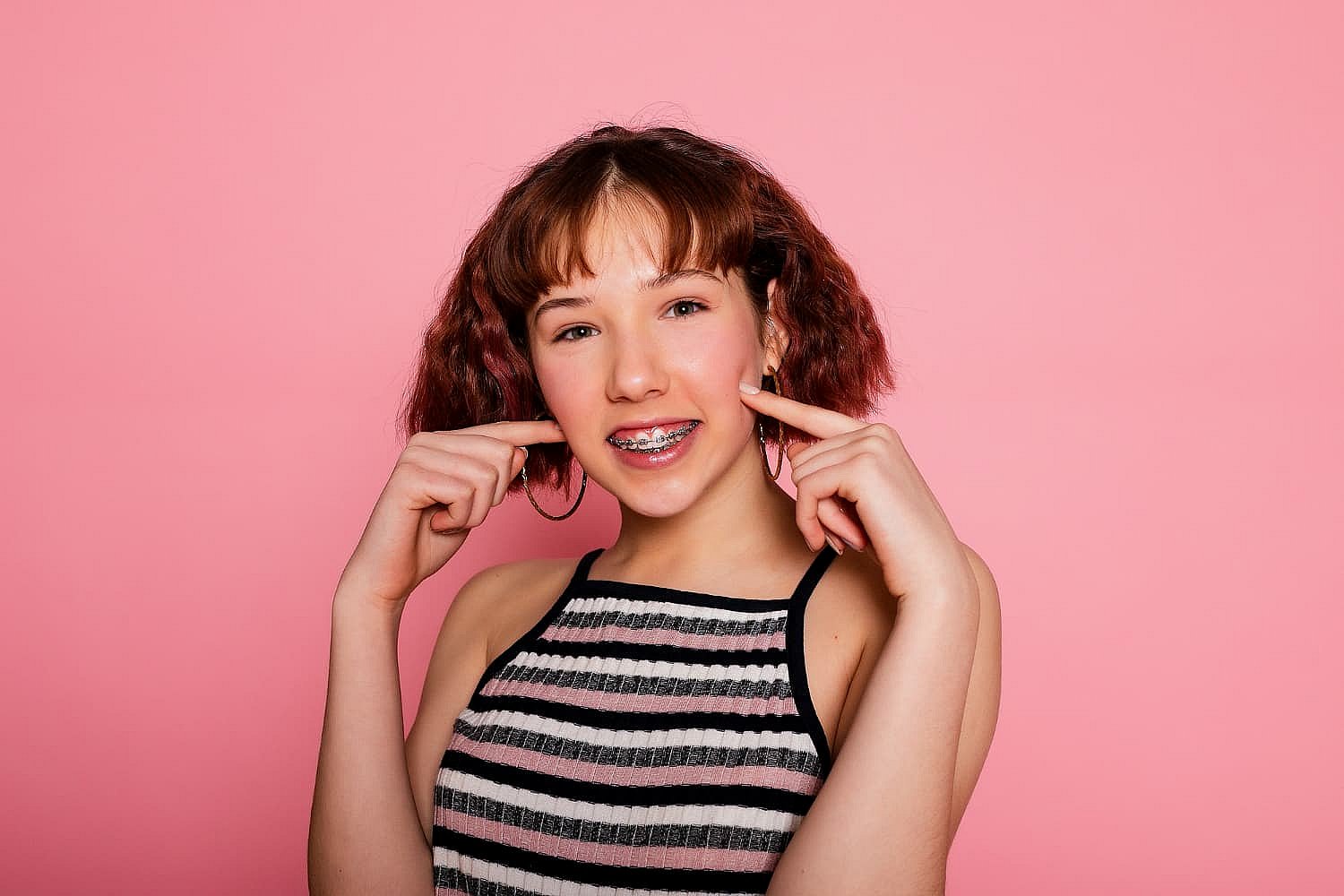 A person with short brown hair smiles, showcasing their braces and pointing at their cheeks. They wear a striped sleeveless top against a pink background, playfully highlighting the debate of Invisalign vs Braces in NYC