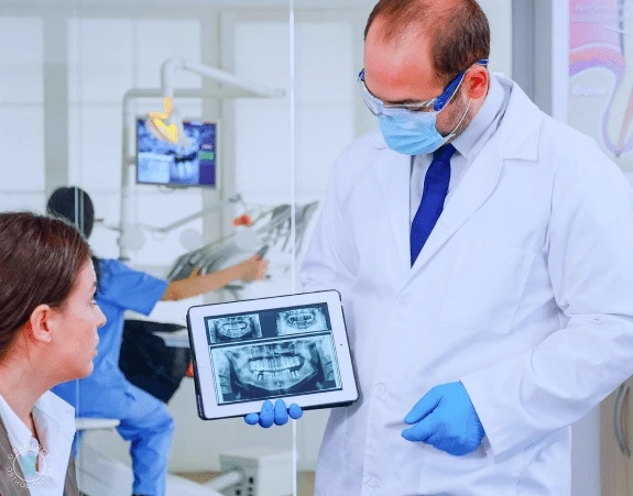 A dentist shows dental X-rays on a tablet to a female patient, discussing potential jaw surgery, while another dentist works diligently in the background in NYC