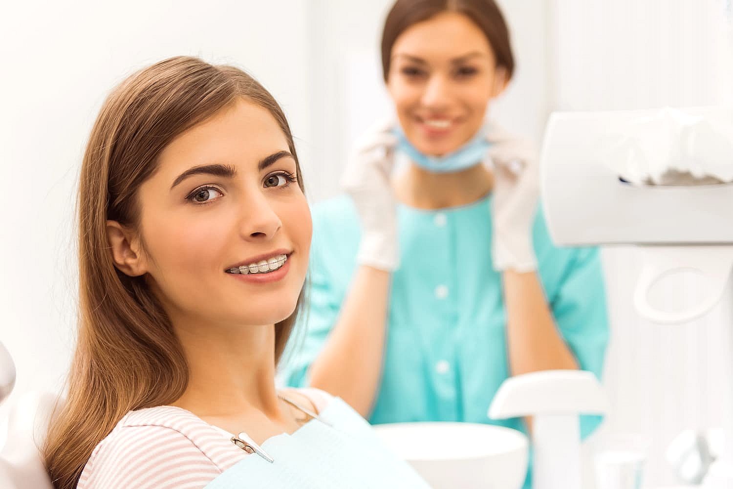 A woman with braces, showcasing adult orthodontics, sits in a dental chair smiling warmly at the camera. In the background, a dental professional in a blue uniform holds a face mask, adding to the atmosphere of care and expertise in NYC