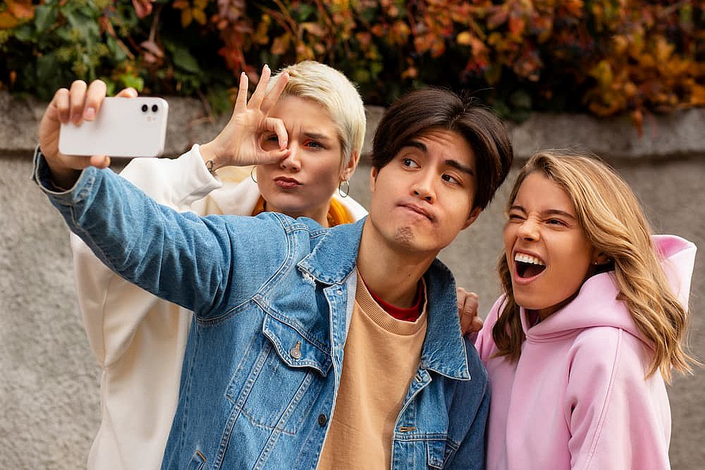 Three friends capturing a selfie outdoors—one in a denim jacket, expertly holding the phone, another playfully gesturing, and the third flashing a wide smile. With the leaves rustling behind them, they could chat endlessly about anything from weekend plans to different types of braces in NYC