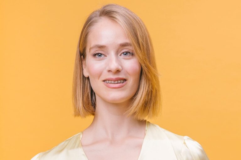 A woman with shoulder-length blond hair and ceramic braces smiles at the camera, set against a solid yellow background in NYC