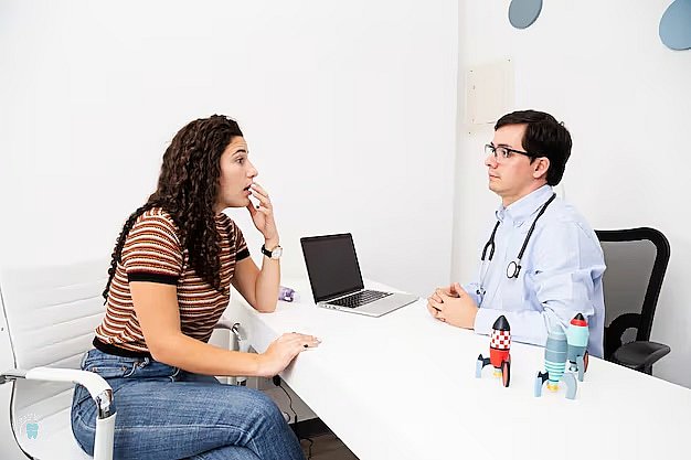 A woman in a striped shirt consults a doctor in an office. The doctor, wearing a stethoscope, discusses tongue thrust concerns while toy rockets and a laptop sit on the desk in NYC